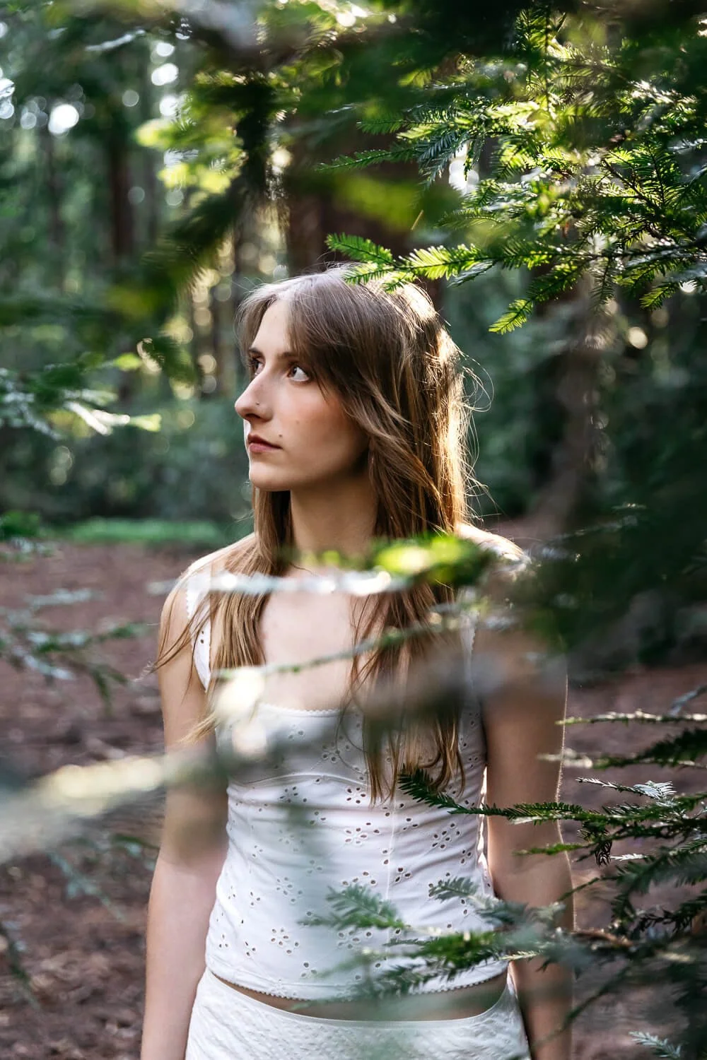 Teen in white skirt and tank top stands amongst redwoods looking contemplative. Shot during senior photo session at Redwood Regional in Oakland.