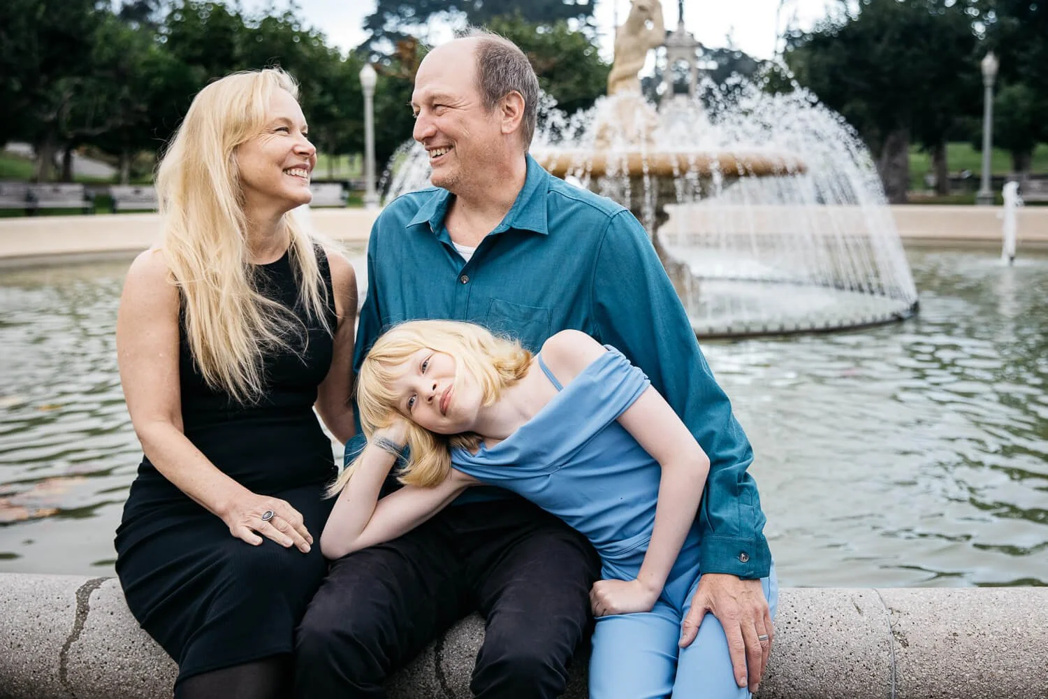 Family of three sitting on the edge of the fountain outside the de Young Museum in San Francisco's Golden Gate Park, mom and dad laughing together while their child leans against dad — San Francisco family mini session.