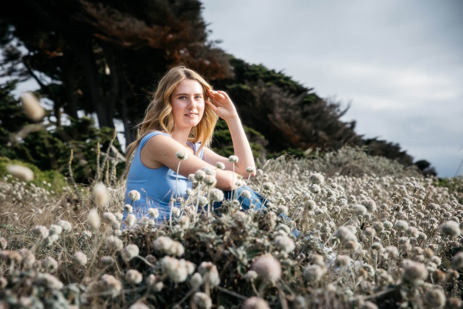 High school senior teen girl with blonde hair in blue shirt sits outdoors in field of white flowers during senior session at Lands End in San Francisco. One hand rests in her hair and she smiles at the camera.
