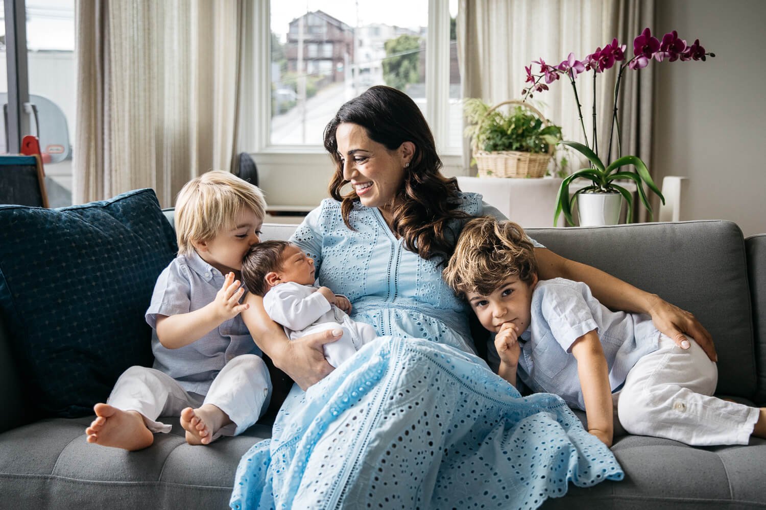 Mama in blue dress holds newborn baby while two little sons cozy up with her on the couch.