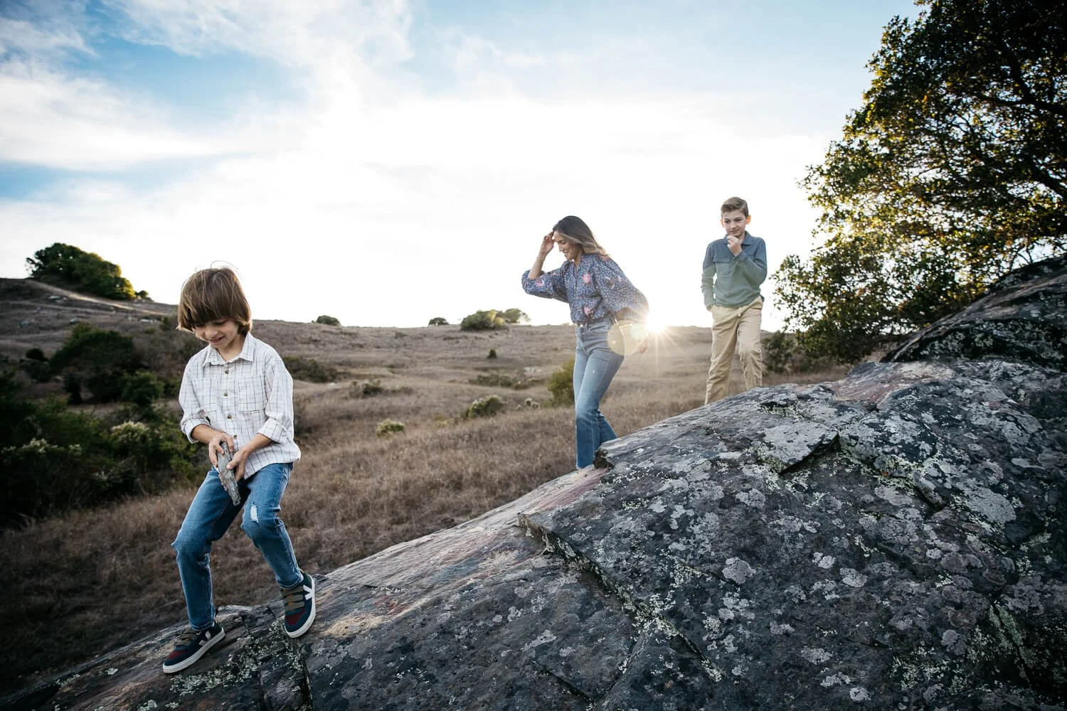 Mom scrambles on large boulder with two young sons, as sun is setting behind them.