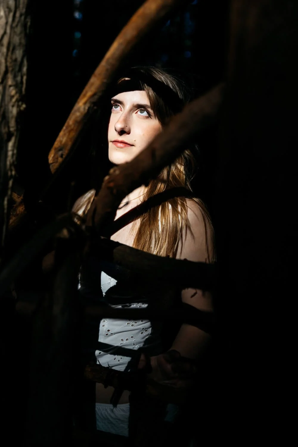 Senior portrait of teen in white tank, standing light tunnel with shadows of branches obscuring part of her body, shot at Redwood Regional in Oakland, CA.