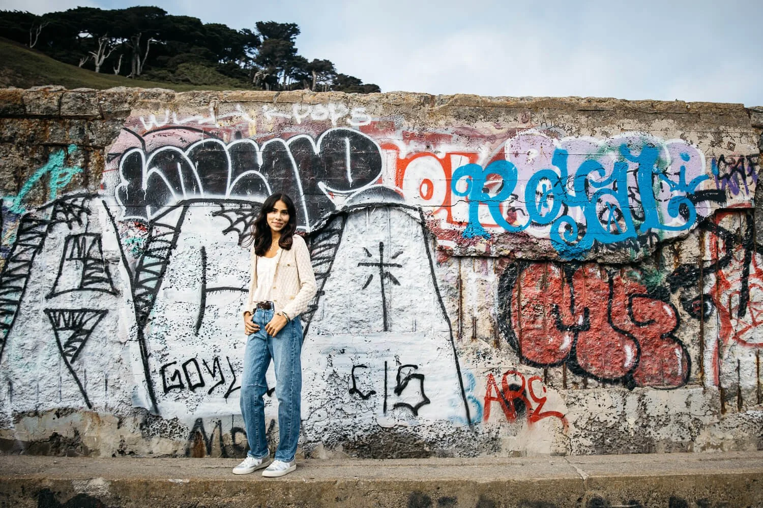 Teen girl in white sweater and jeans stands in front of graffiti mural at Sutro Baths.