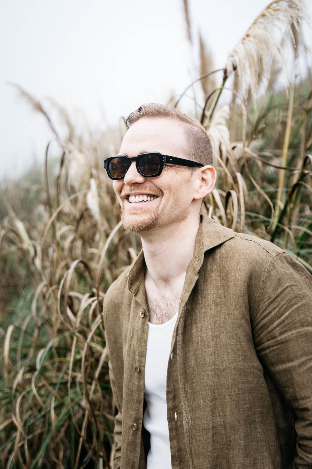 Man in open linen shirt and sunglasses, looks off in distance, smiling with tall grass behind him.