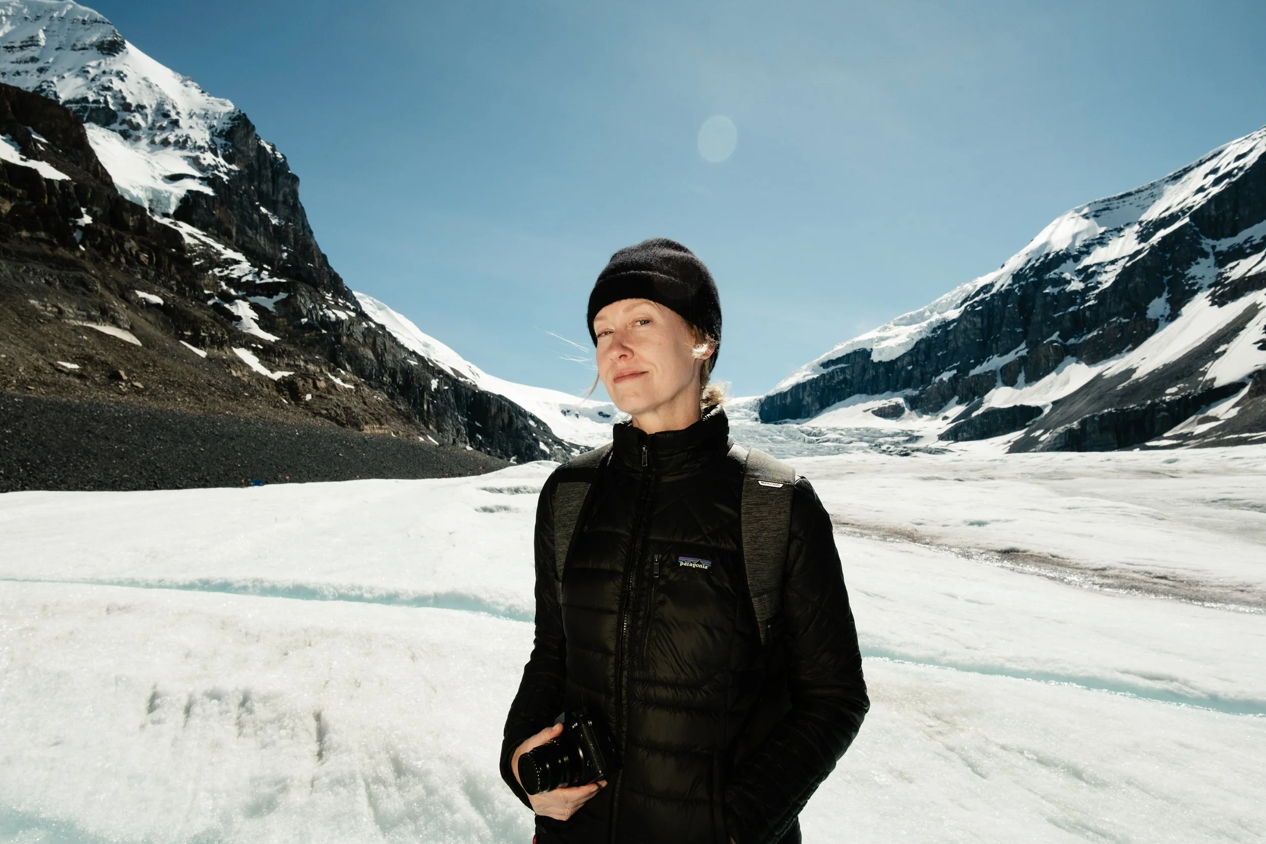 Woman holding camera and wearing all black stands on icy mountain and smiles