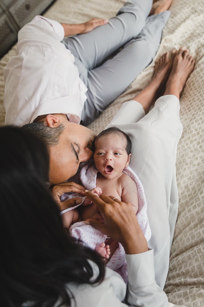 Newborn lying on mom's legs coos while Dad reclines nearby and kisses baby's cheek.