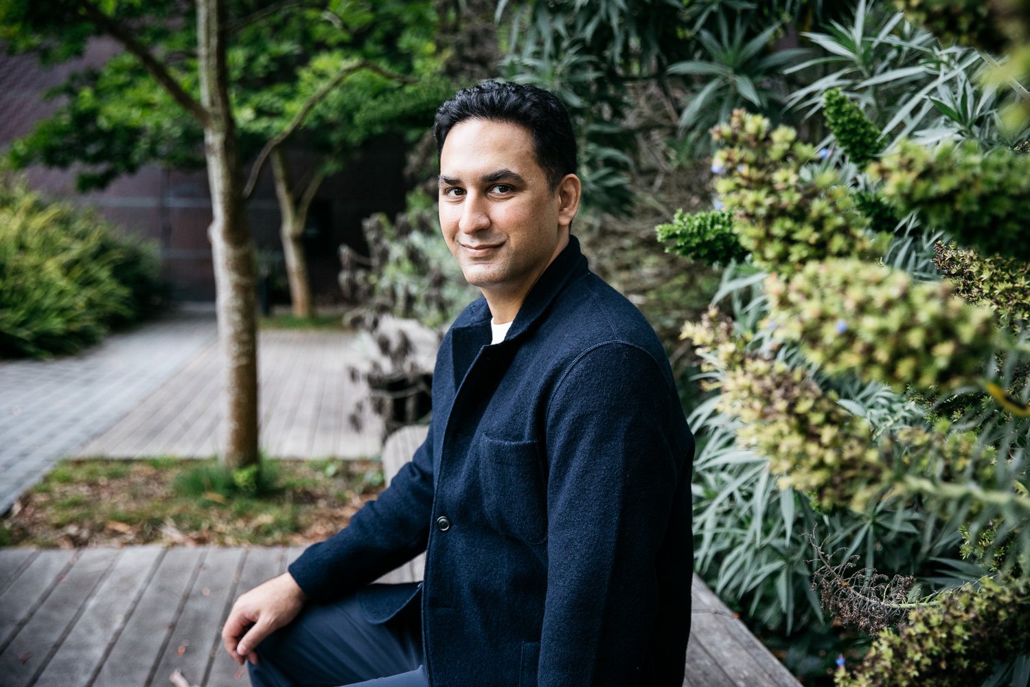 Man with dark hair and navy wool jacket sits amongst California landscaping during outdoor dating app portrait photo shoot in San Francisco.