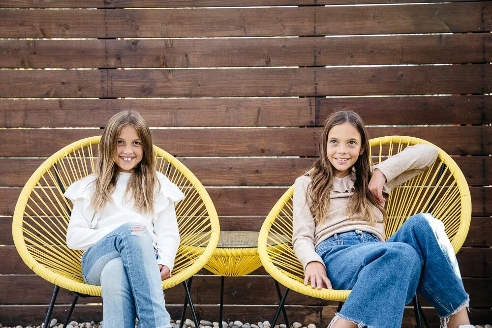Two tween sisters sit in yellow chairs in front of a wooden fence in the backyard of their Lafayette home. They are both looking at the camera and smiling. Taken during a lifestyle family photo session.
