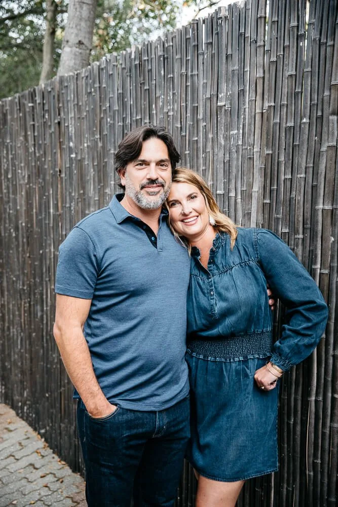 Family portrait of a mother and father leaning into each other in the backyard of their home in Lafayette. They both smile at the camera, with one arm around each other and the other in their respective pockets.