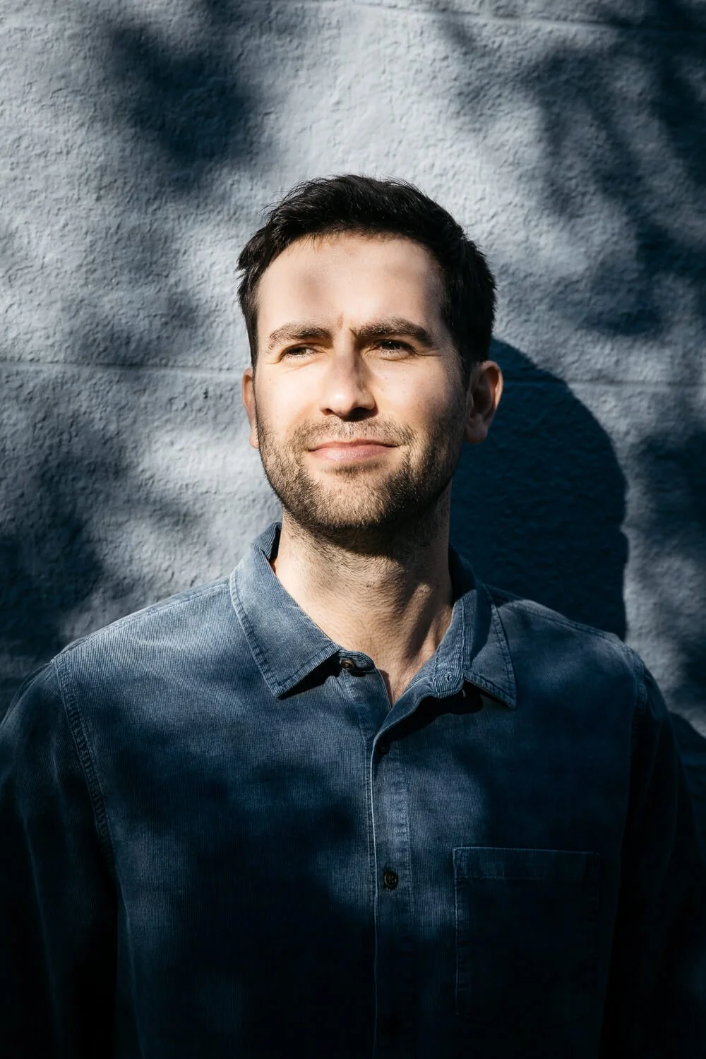 Man in blue shirt stands against blue wall in dappled sunlight in San Francisco.