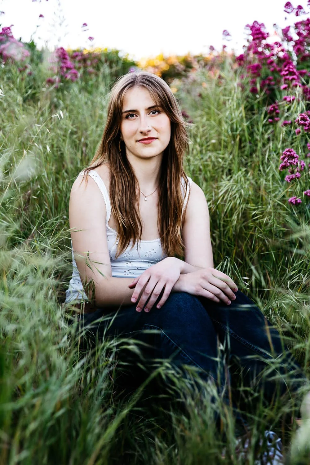 Berkeley senior photographer captures high school senior with long auburn hair sitting among tall grass and pink wildflowers at the Albany Bulb, wearing a white eyelet tank top and dark jeans.