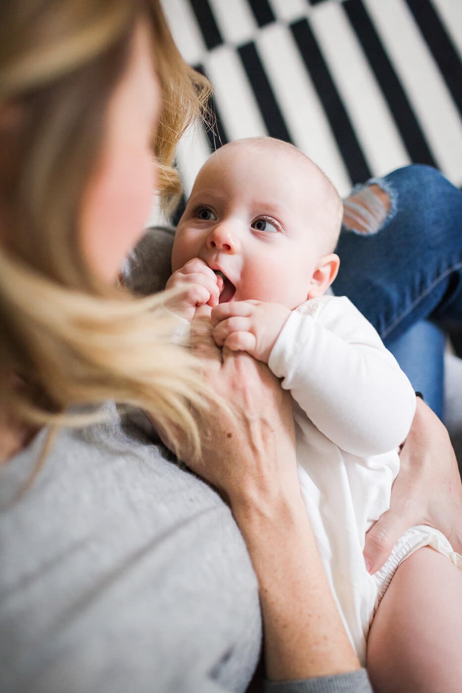 Mom gazes down at smiling baby in white onesie against a black and white striped rug.