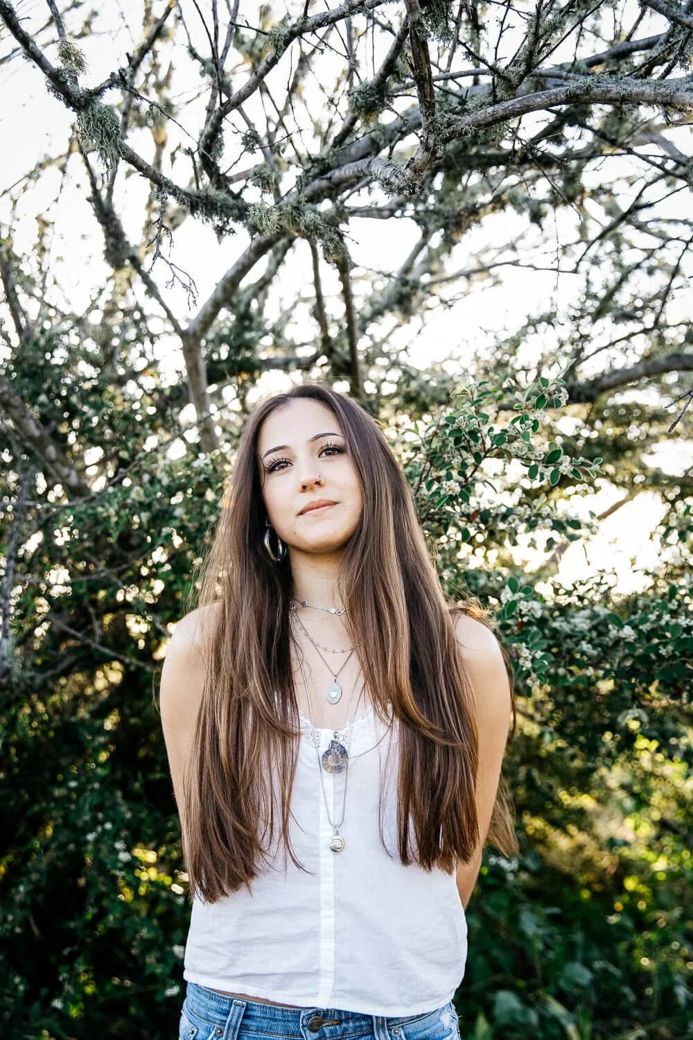 San Francisco senior photographer captures high school senior with long brown hair standing beneath a lichen-covered tree at the Albany Bulb, wearing a white tank top and layered silver necklaces.