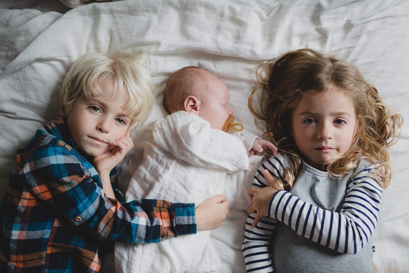Brother and sister lie on bed with white spread and hug their sleeping newborn brother.