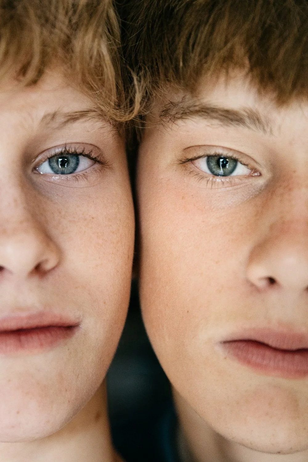 Close-up of two teen boys' faces pressed cheek-to-cheek and gazing at the camera, cropped to show one half of each of their faces.
