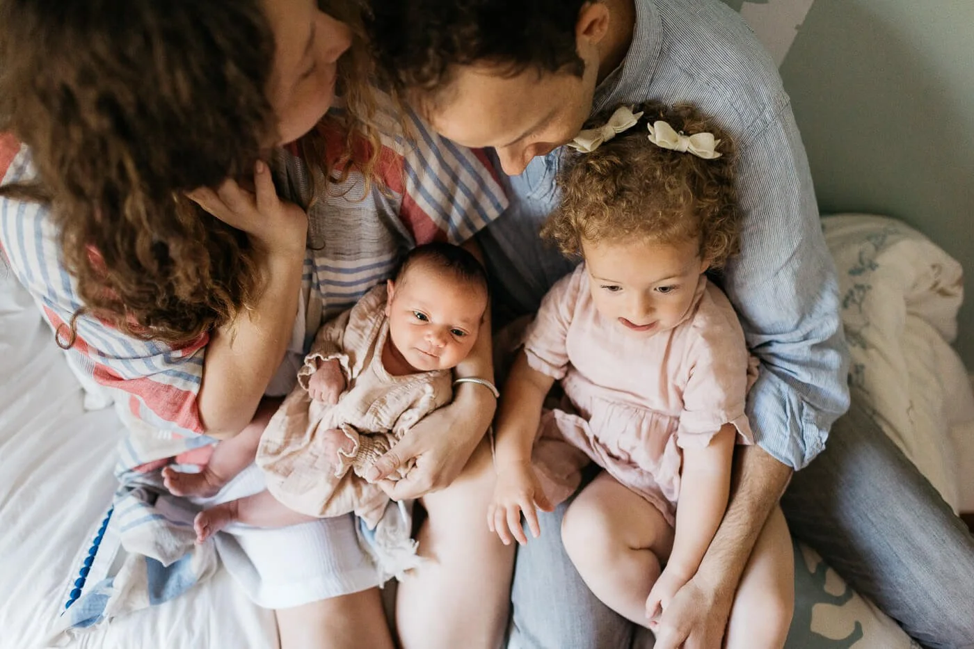 Family snuggle overhead shot with toddler and newborn daughters wearing soft pink dresses, sitting in mom and dad’s lap. Taken during newborn lifestyle session at home in Palo Alto.