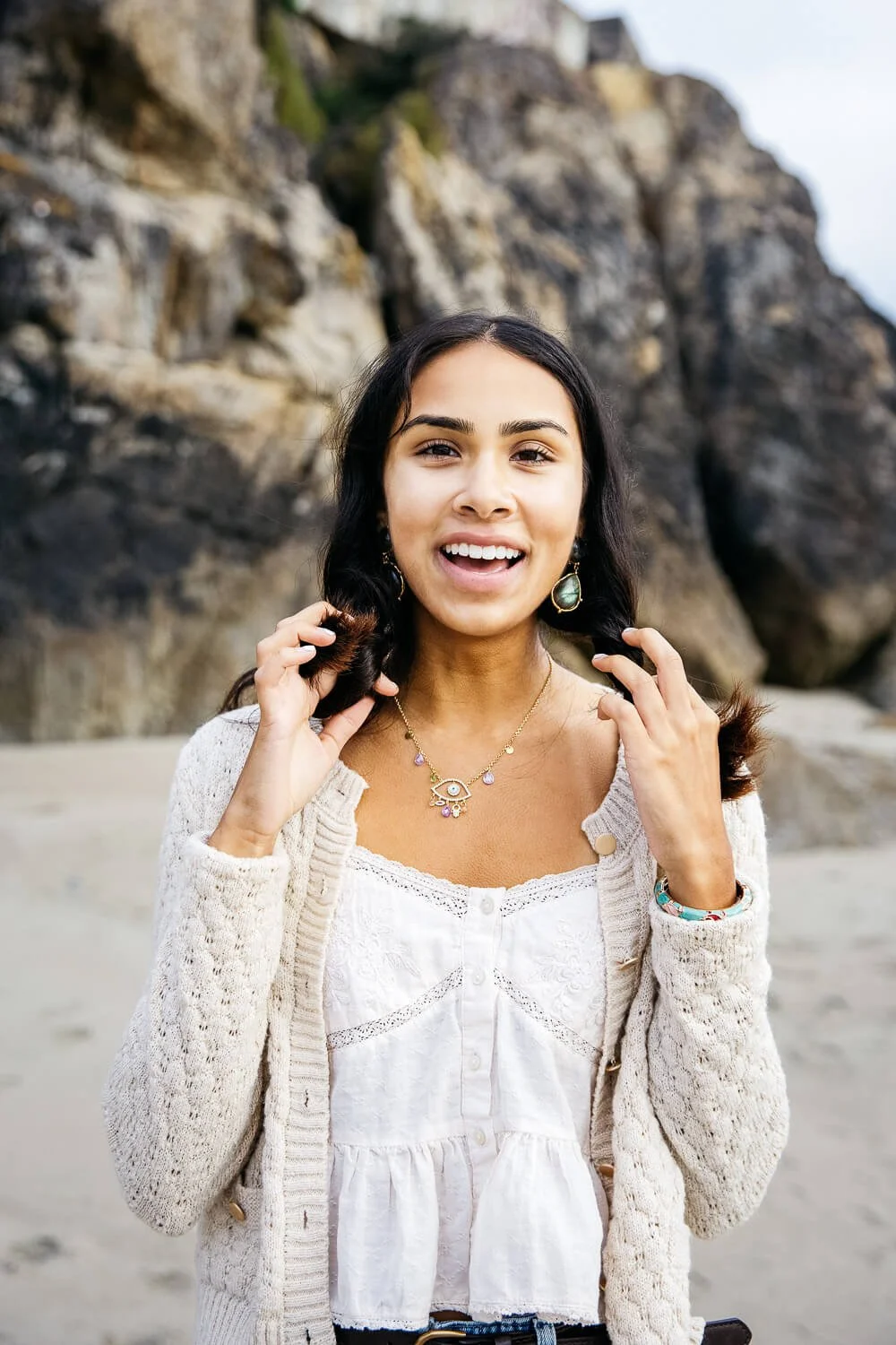 South Asian girl in cream sweater and jeans stands on beach, playing with hair, shot during senior photo shoot at Ocean Beach in San Francisco.