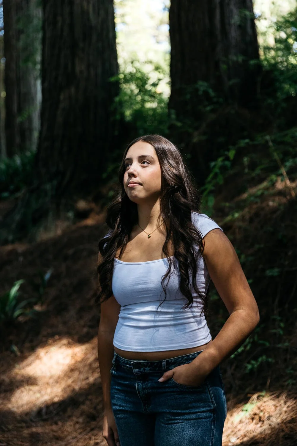 Teens with long brown hair and wearing white tank and jeans, stands in shaft of sunlight in a redwood grove during senior portrait session Blake Gardens.