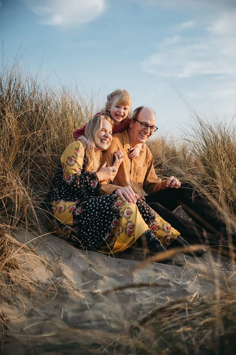 Toddler girl squeeze hugs mom and dad as they sit the dunes at Ocean Beach in San Francisco, as the sunset light creates warm glow.