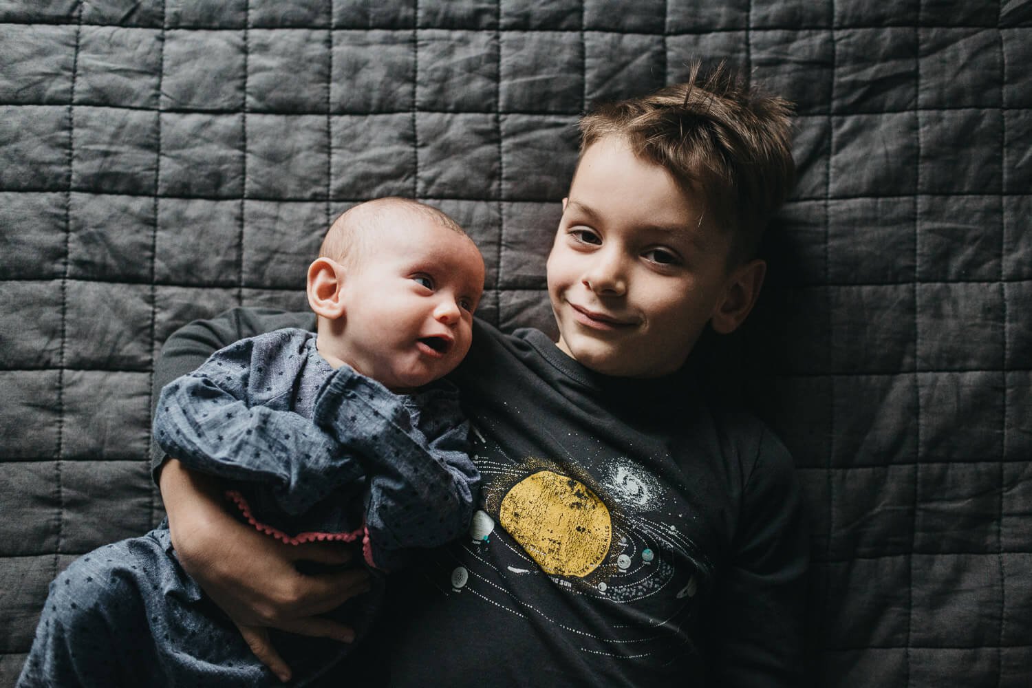 Young boy lays on grey comforter and holds newborn sibling who gazes at him during a newborn session in San Francisco.