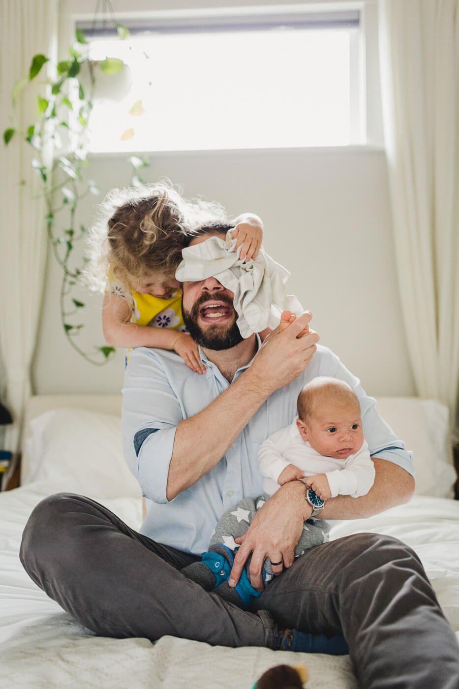 Dad holds newborn while toddler climbs on his back and covers his face with blanket, taken at home in Berkeley.