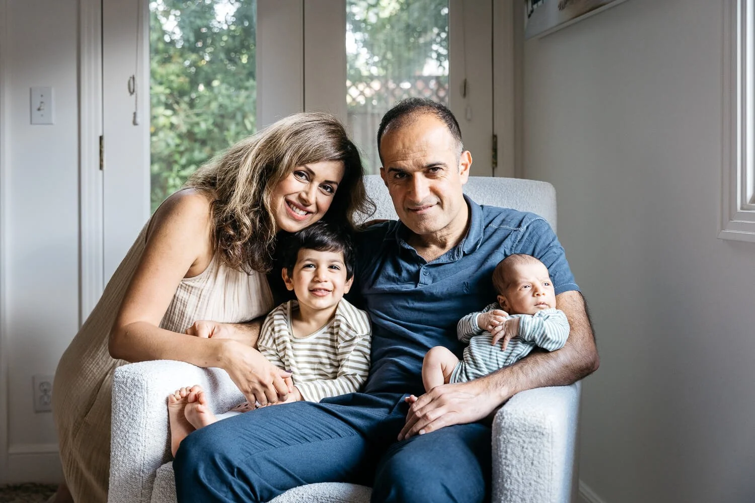 A family of 4 snuggles together in living room, newborn baby and toddler brother on dad's lap in a white chair. Mom kneels next to them in leans in. Everyone smiles at the camera.