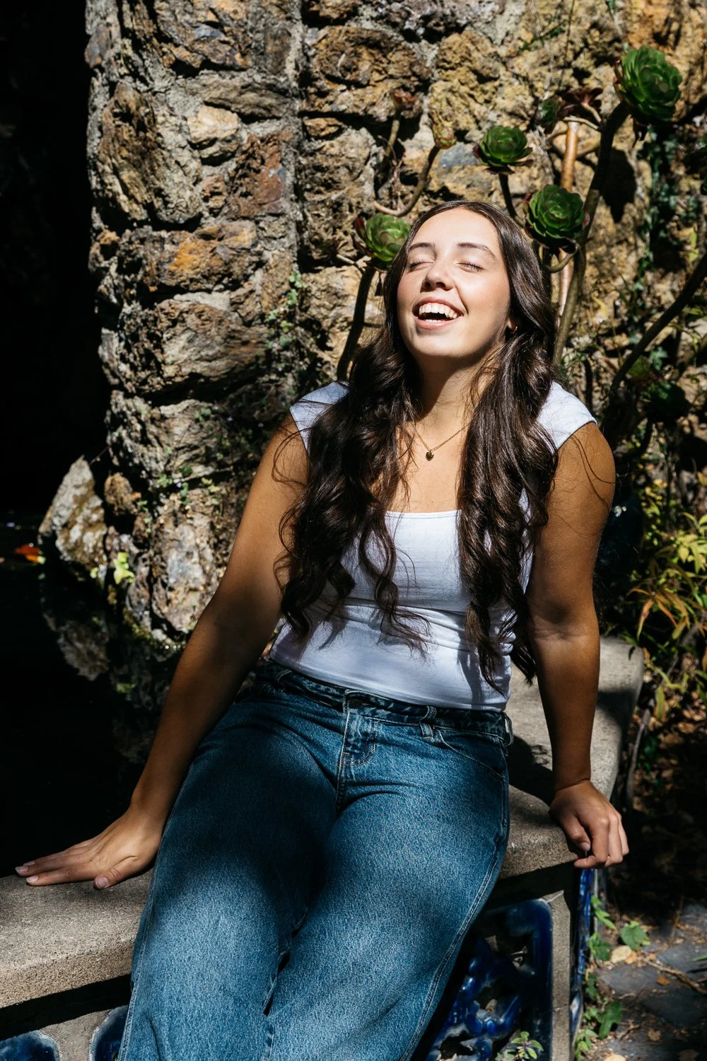 High school senior girl in white tank and jeans sits on stone bench and laughs naturally with closed eyes as the sun shines down on her. Shot during senior picture session at Blake Garden in Berkeley.