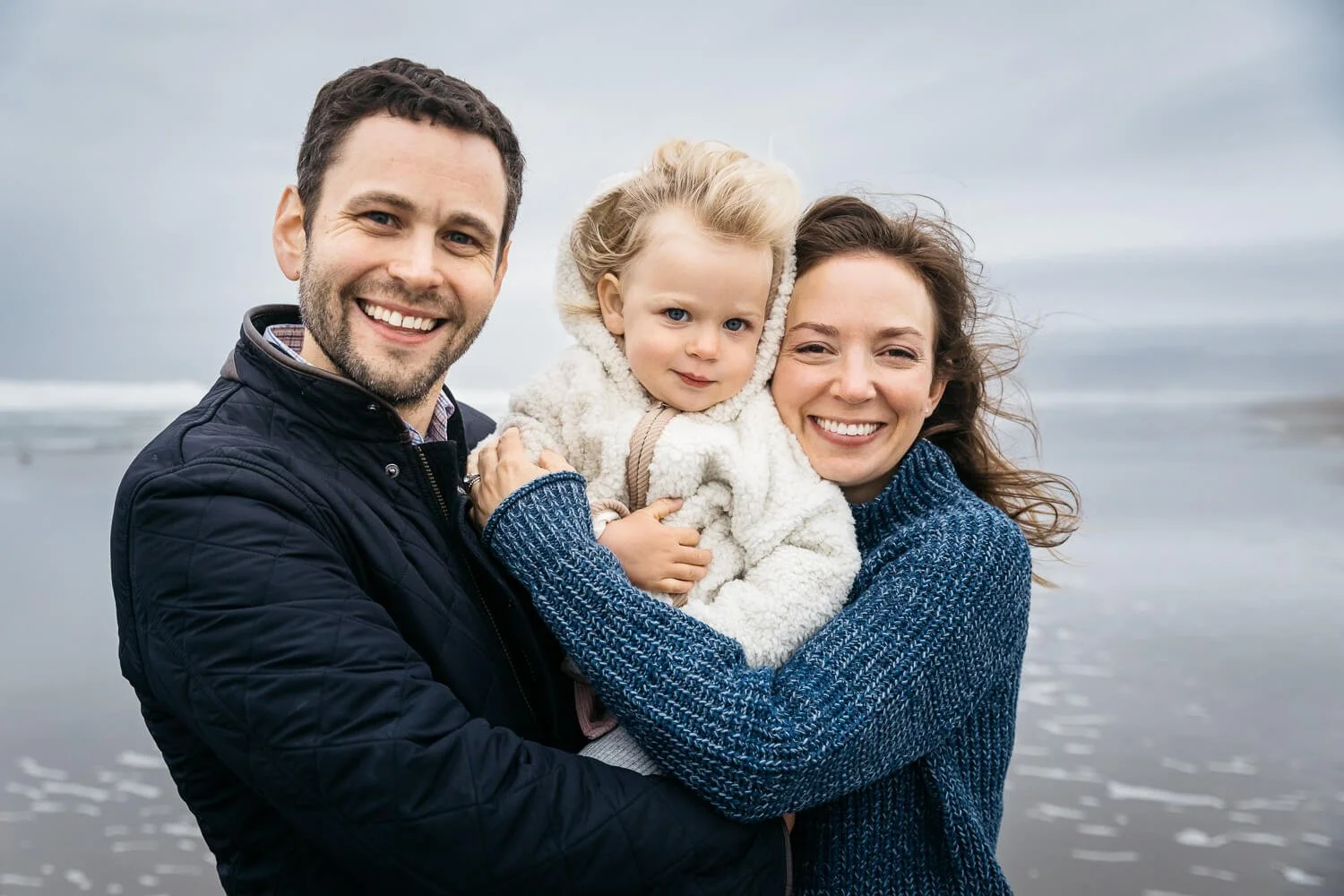 A family of 3 stand on a windy Ocean Beach together, snuggled up and hugging. The toddler is snuggle dbetween mom and dad, and the wind blows through everyone's hair. Waves roll in behind them. The family smiles at the camera.