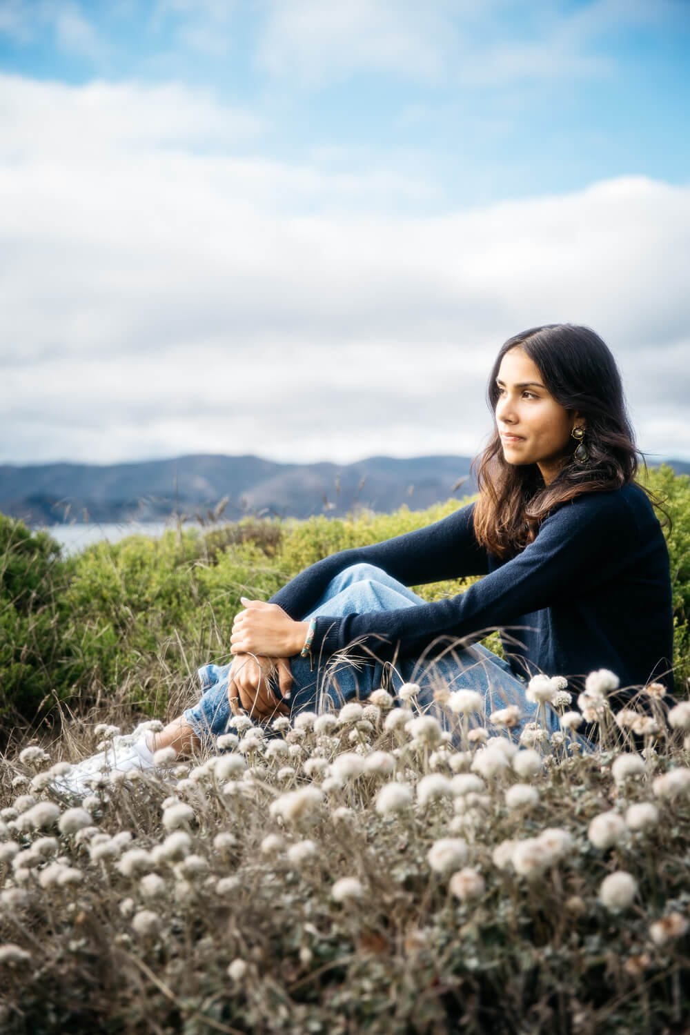 Profile view of south Asian girl in navy sweater and jeans sitting among coastal scrub plants at Lands End, shot during senior photo shoot in San Francisco.