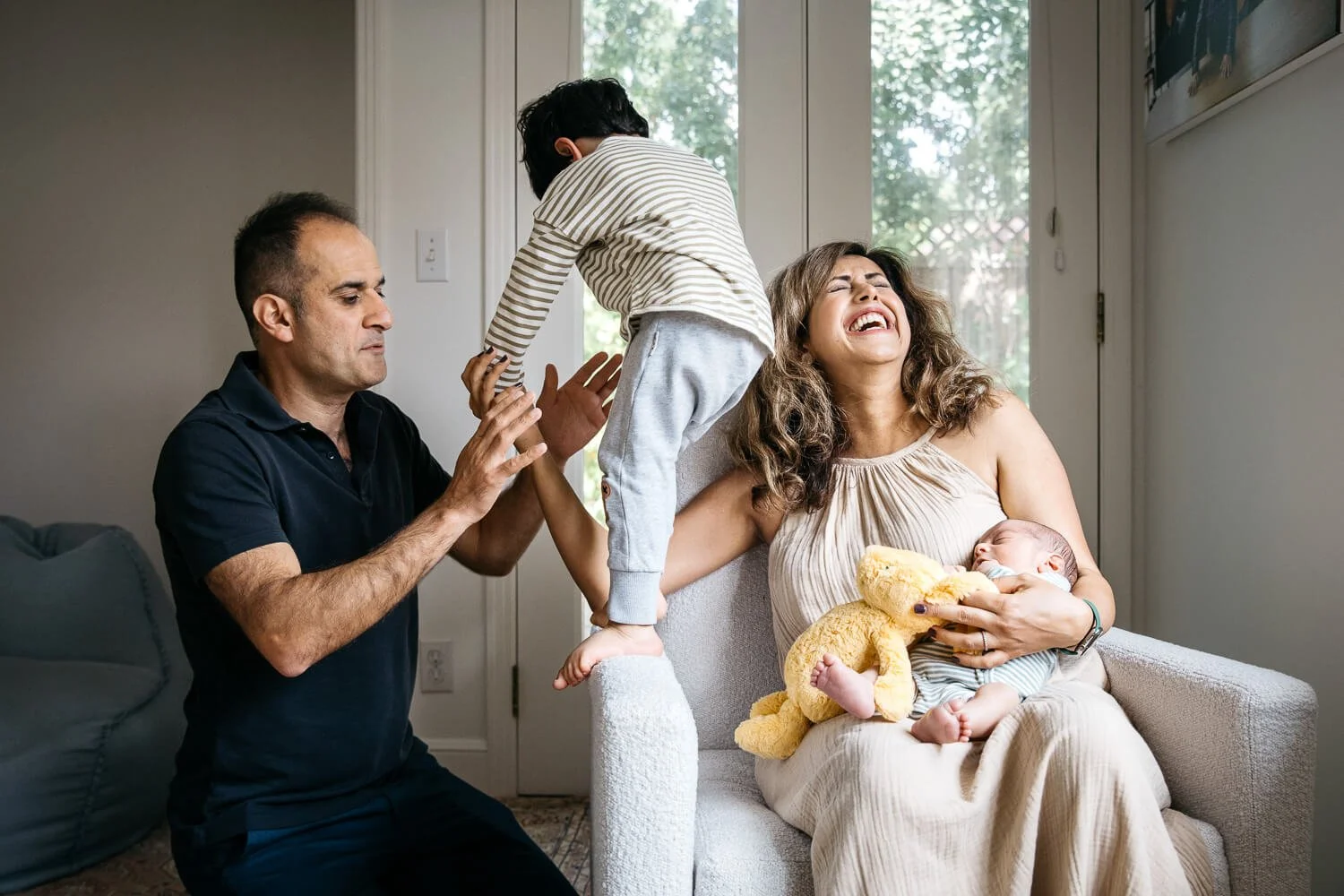 Toddler brother climbing on armchair while dad spots him, mom laughing while holding sleeping newborn baby. Taken during in home newborn lifestyle session in San Francisco.