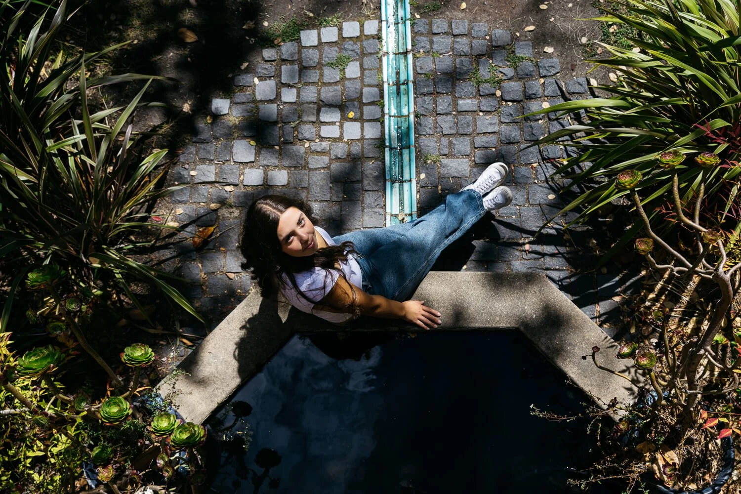 Overhead shot of teen girl in white tank and jeans sitting at a fountain, shot during senior portrait session at Blake Garden in Kensington.