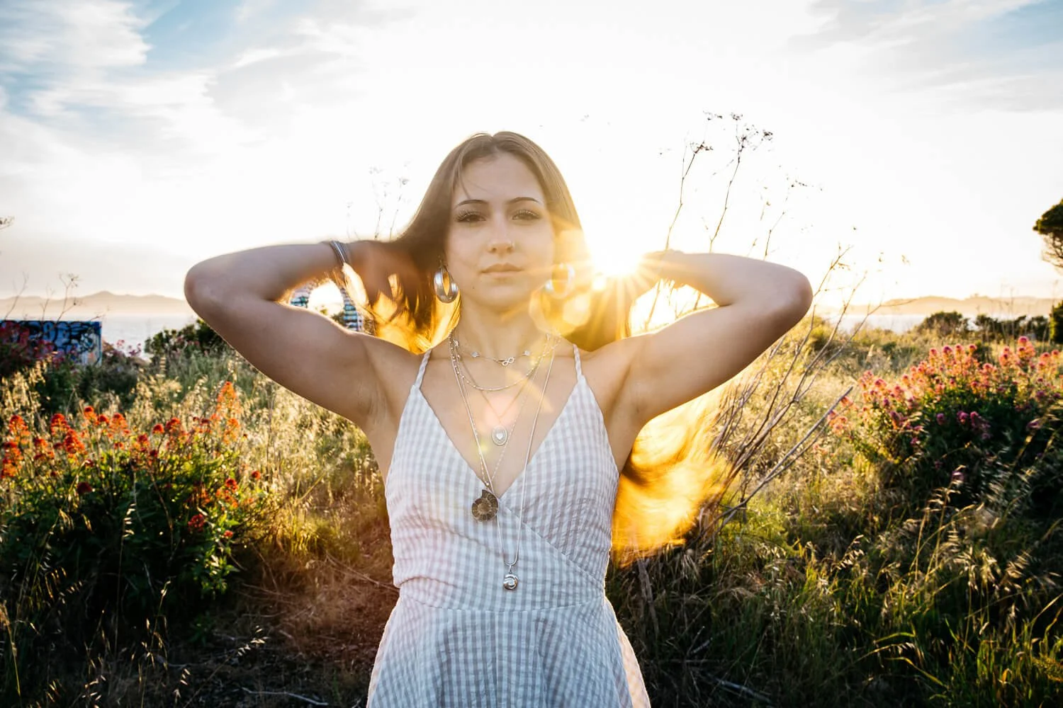 Teen girl in long gingham dress stands with sunset light behind her and wind blowing her hair, shot during senior photo session at Albany Bulb.