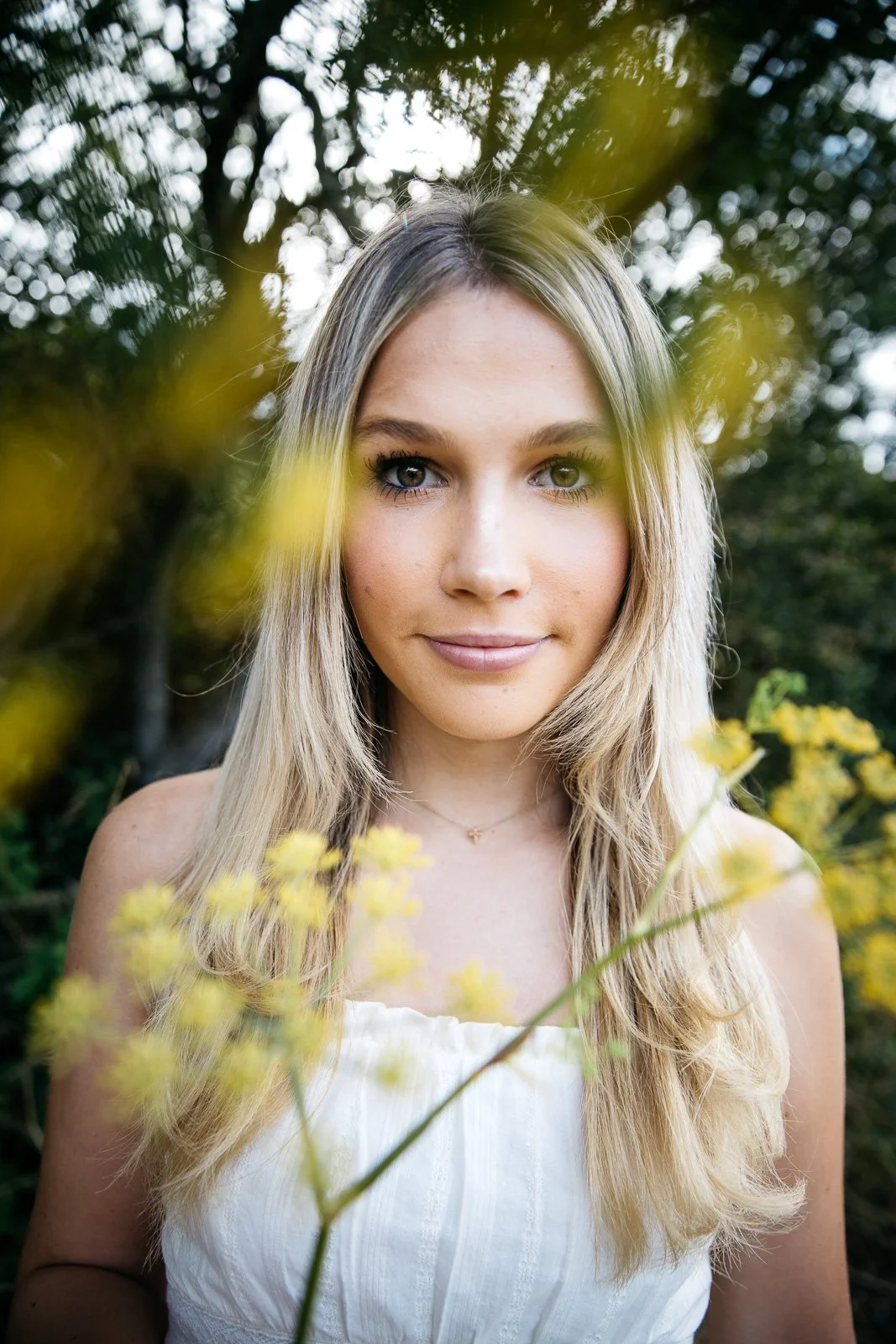 Teen girl in white tank stands in patch of yellow flowers and looks through them at camera, shot during senior session in the Marin Headlands.