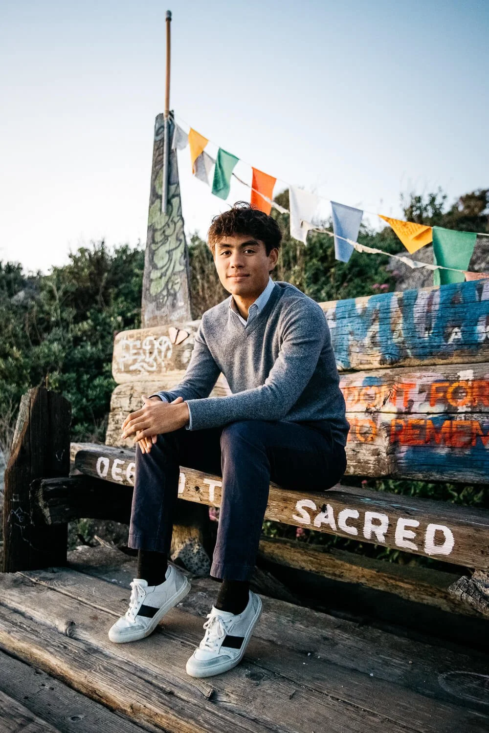 Teen boy sits on bench with painted flags behind him, shot at the Albany Bulb at sunset.