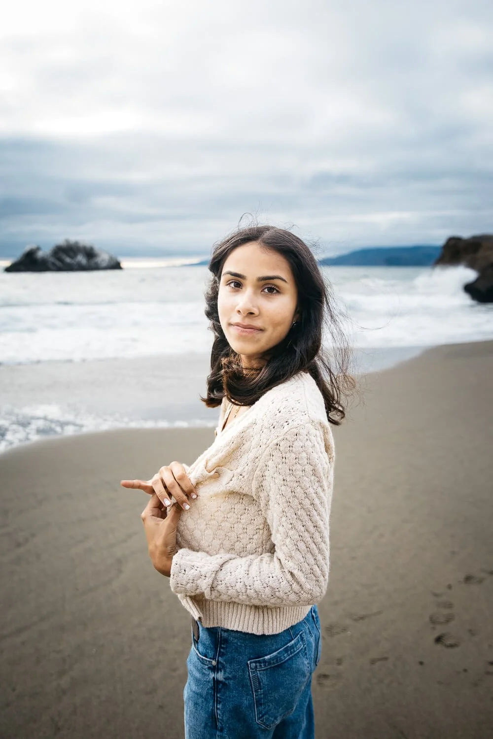 South Asian girl in cream sweater and jeans stands on beach, holding sweater closed, shot during senior photo shoot at Ocean Beach in San Francisco.