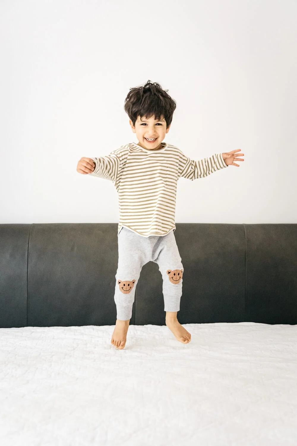 A toddler boy jumps on his parents bed, arms up and a large smile.