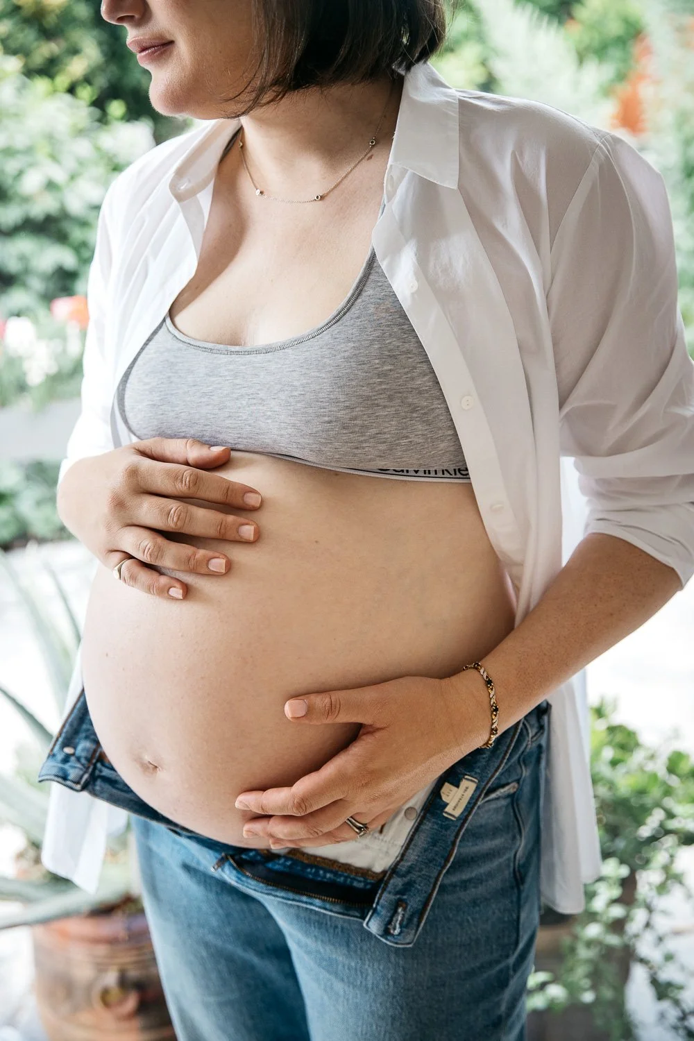 Crop of woman's pregnant belly, wearing open white cotton shirt during maternity photo session in Palo Alto.