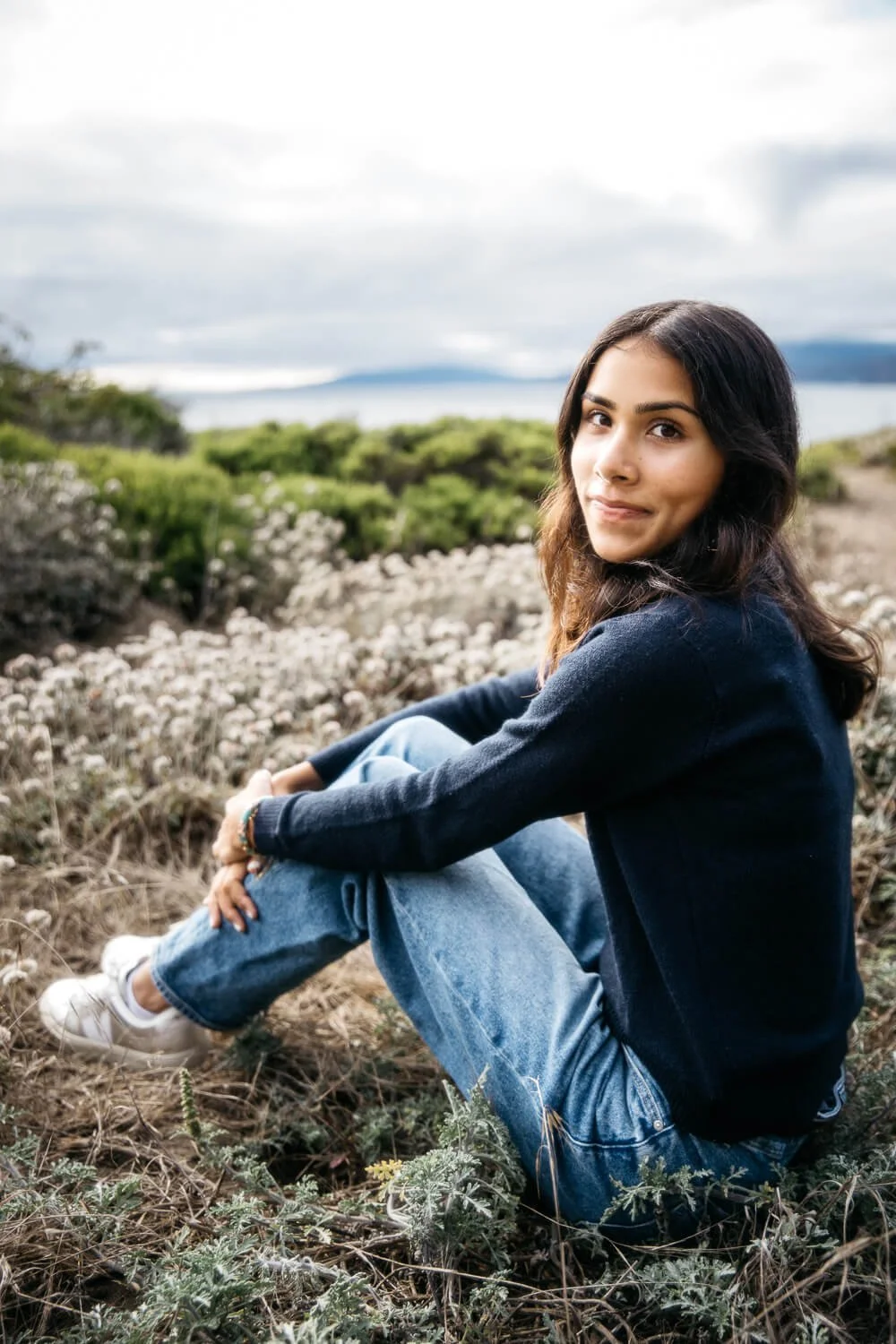 South Asian girl in navy sweater and jeans sits among coastal scrub plants at Lands End, shot during senior photo shoot in San Francisco.