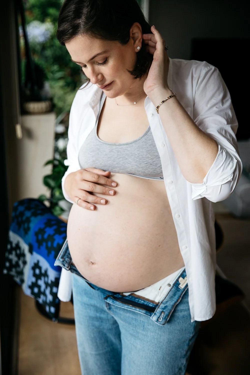 Pregnant mama wearing open white cotton shirt exposes pregnant belly and gazes down, one hand on top of her baby bump. Shot during a lifestyle maternity session at home in San Francisco.