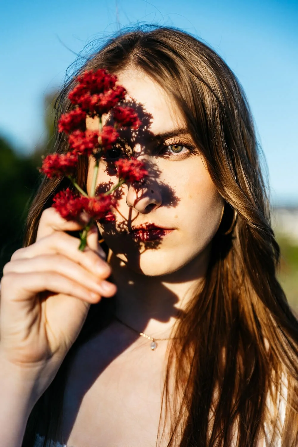 Teen high school senior girl holds fuscia flower in front of her as sun casts shadow that partially obscures her face. Shot during senior photo session at Albany Bulb.