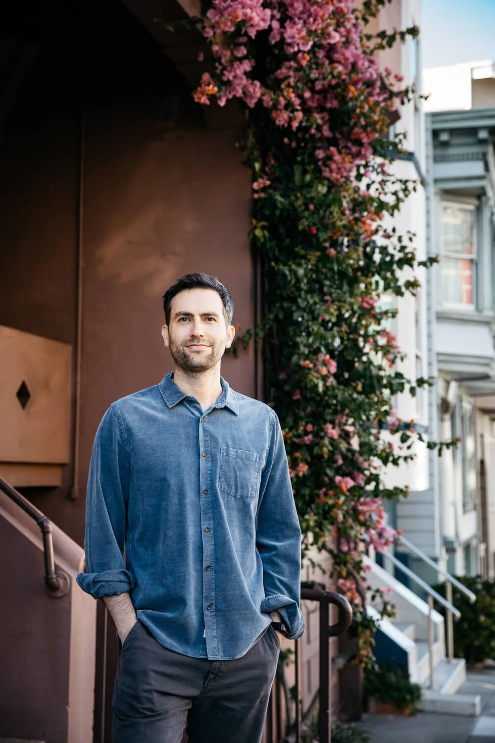 Man in blue shirts stands in front apartment building on Lombard street with bougainvillea growing up the side.