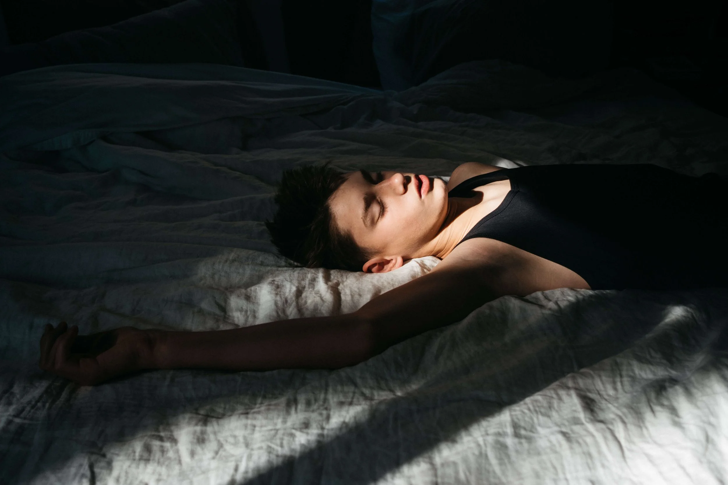 Shadowy room with one beam of sunlight coming through and lighting up the face of a teen boy lying on the bed.