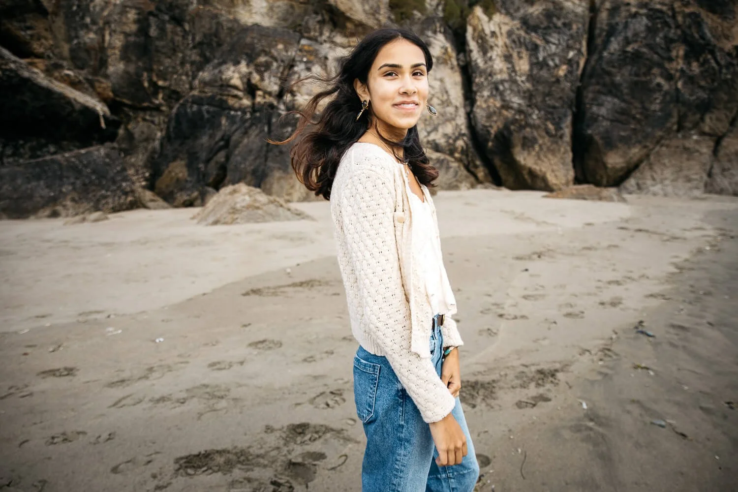 High school senior young woman in white sweater and jeans walks in the sand in front of cliffs at Ocean Beach in San Francisco. The wind blows through her hair as she smiles.