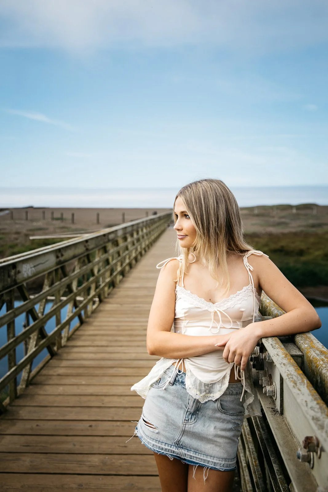 Teen girl with blond hair, white satin tank, and short denim skirt stands on footbridge at Rodeo Beach, during senior photo session.