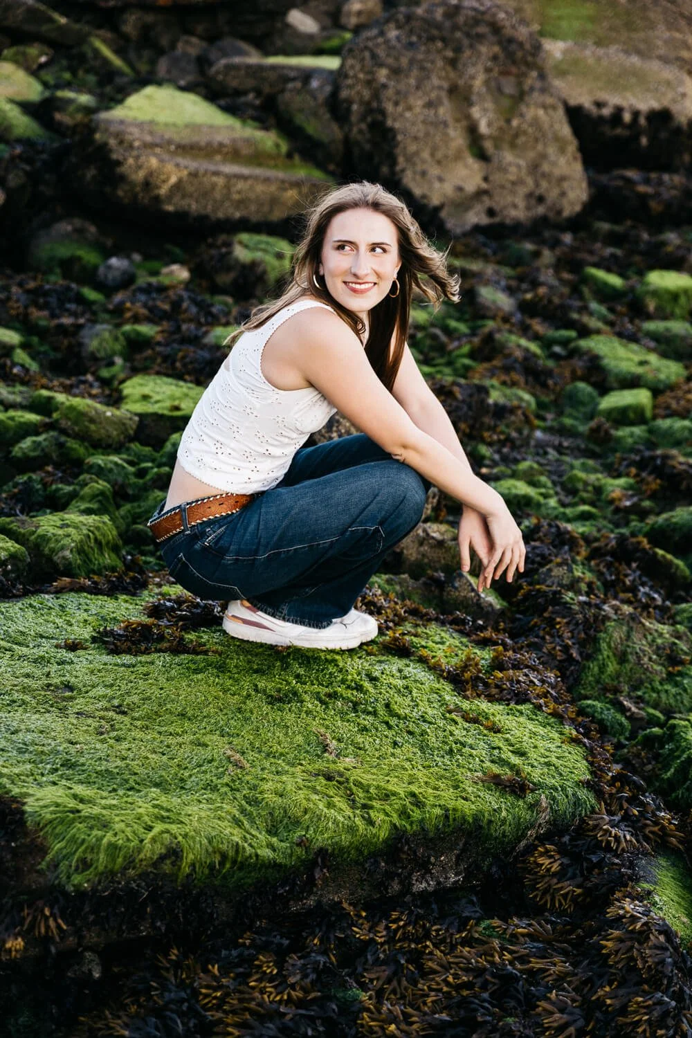 Berkeley senior photographer captures high school senior crouching on moss-covered rocks at the Albany Bulb shoreline, smiling over her shoulder in a white eyelet tank top and dark jeans.