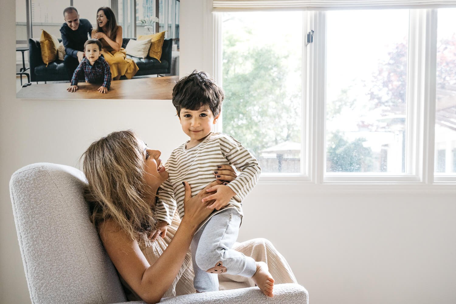 Mom lifting and laughing with her toddler son in a sunlit armchair during in home newborn lifestyle session in San Francisco. On the wall behind them is a framed photo of the family taken in years prior.