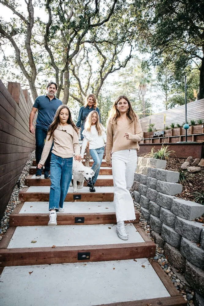 Family of five, with three tween girls, walk down stone steps with white labrador running down between them.