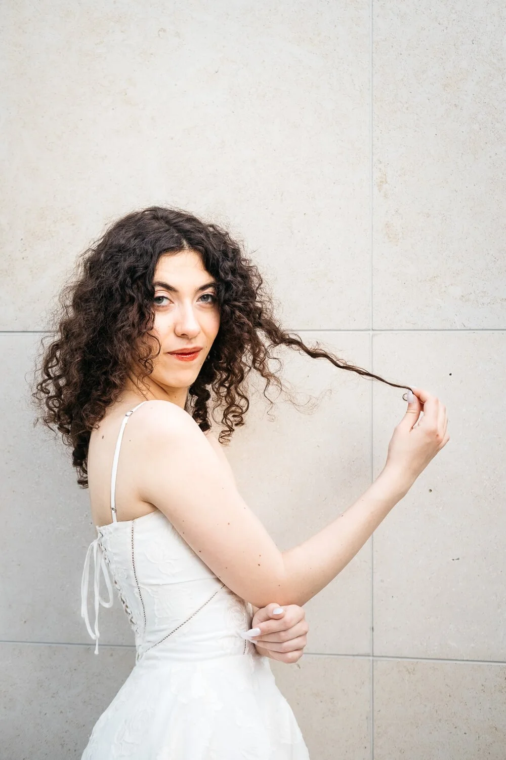 Young woman with dark curly hair pulls her curls, while wearing white dress and standing in front of a while marble wall.