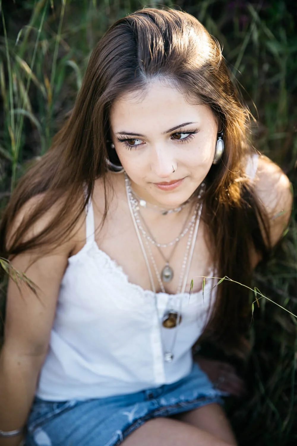 San Francisco senior photographer close-up portrait of a high school senior with long brown hair sitting in tall grass at the Albany Bulb, wearing a white lace tank and layered silver necklaces.