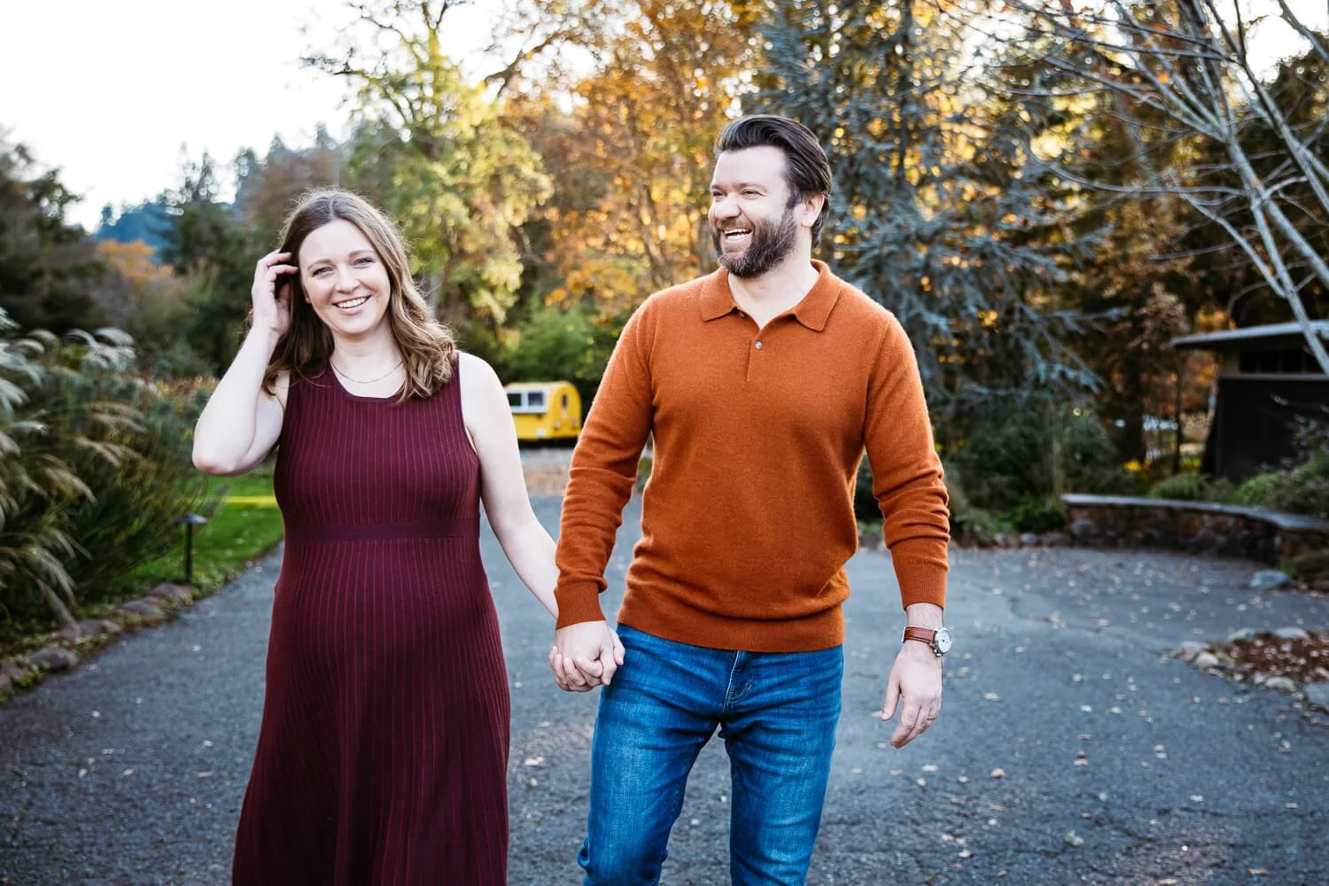 Pregnant mama in maroon dress walks holding hands with husband, wearing brick orange sweater. They both laugh and fall colors in the trees are behind them.